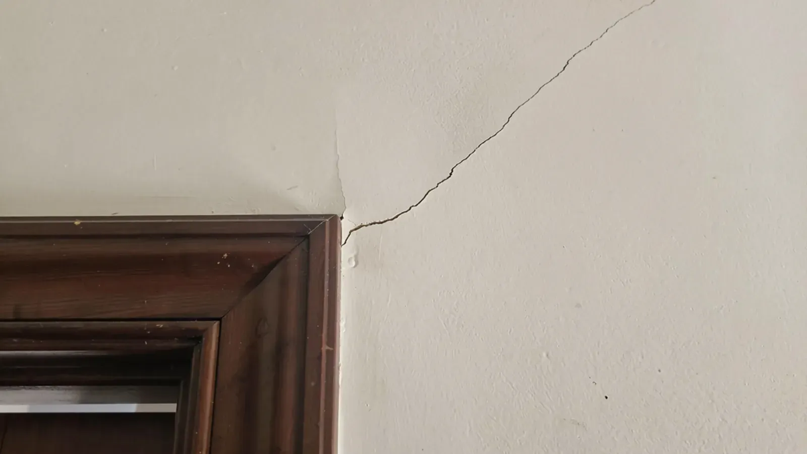 A fine hairline crack running diagonally across a painted interior wall from the corner of a dark-wood door casing in an older home