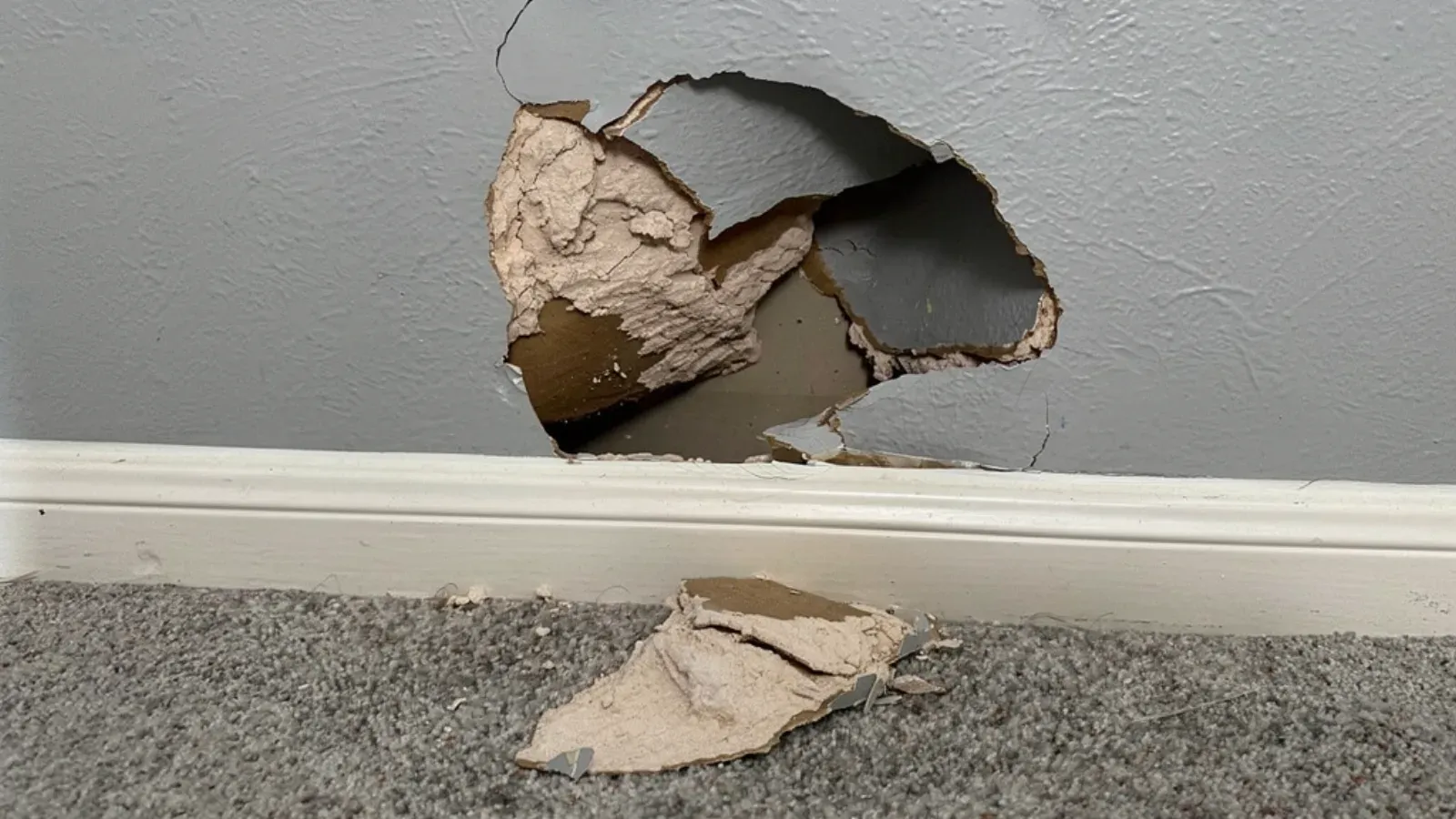 Large impact hole low on an interior drywall wall above the baseboard, a broken piece of drywall on the floor below