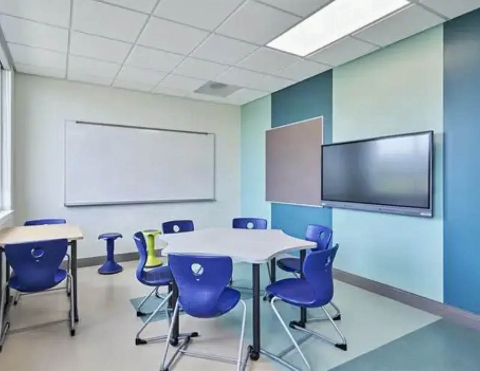 Small breakout classroom with finished painted drywall walls in blue and teal, a suspended ceiling, a whiteboard, a wall-mounted display screen, and a group table with molded chairs