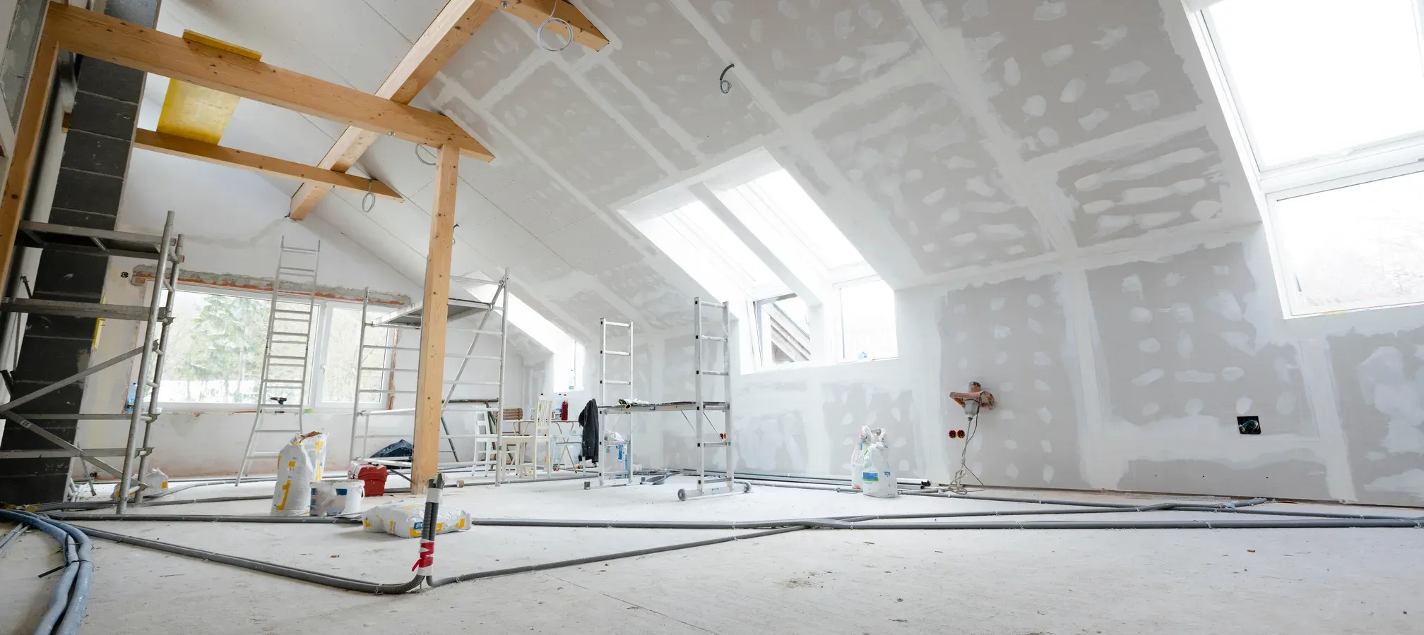 Wide attic loft under a sloped drywall ceiling with skylights, mobile scaffolding, and taped-and-mudded walls mid-finish, with materials staged on the bare floor