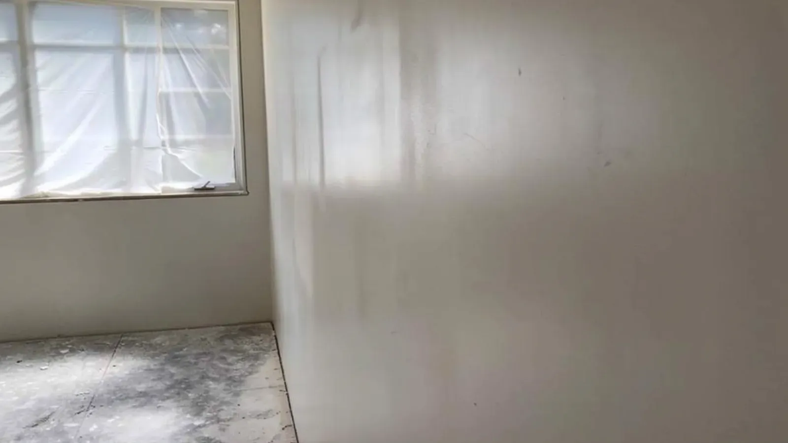 A freshly skim-coated interior wall running back from a plastic-masked window, the surface an even off-white with no visible seams
