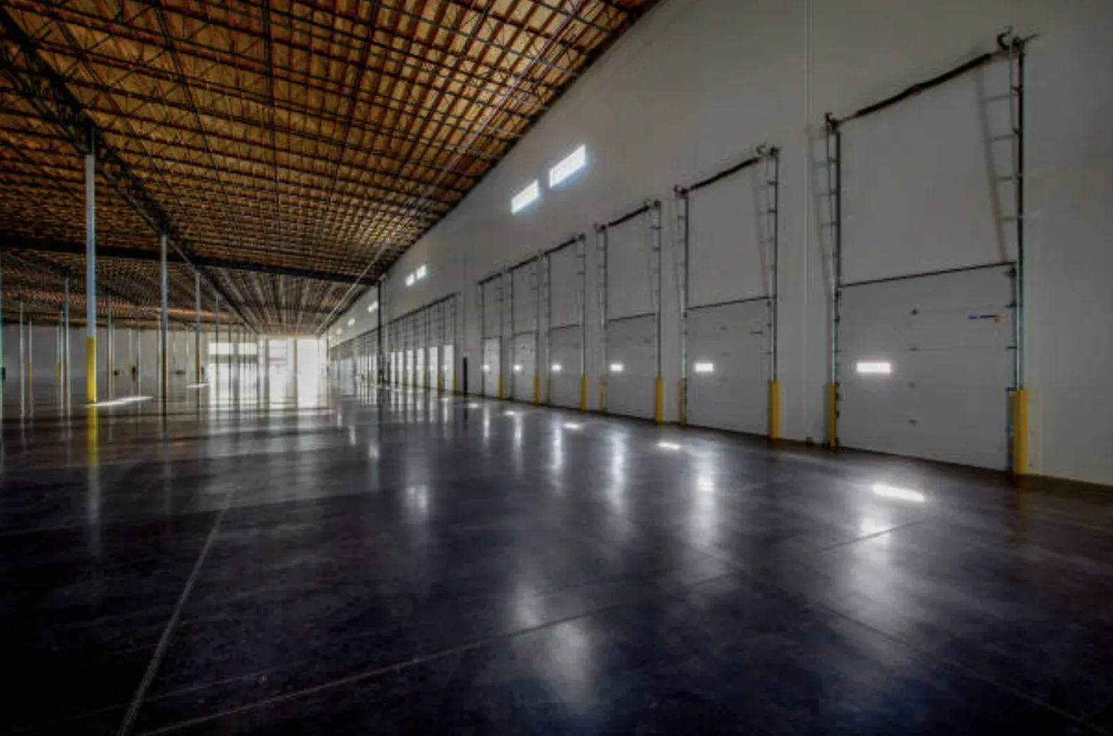 Empty industrial warehouse with a long run of finished white wall panels, loading-dock doors with yellow bollards, and a polished dark concrete floor under a timber roof deck